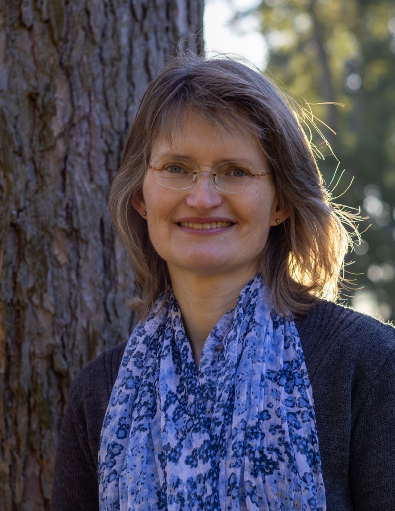 Zu sehen ist eine Portrait Aufnahme von Vera Reimann.
Sie lehnt an einem Baum und lächelt in die Kamera. Sie hat mittellange blonde Haare, blaue Augen, trägt eine rahmenlose Brille, einen grauen Pullover und ein hellblauen Schal.